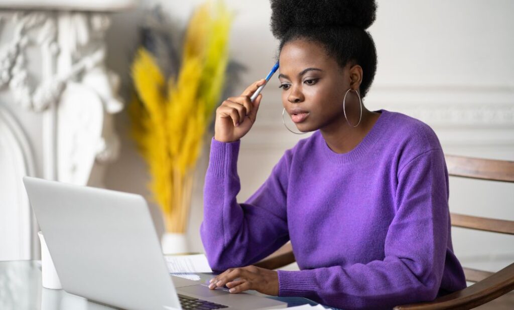 A LADY HOLDING A PEN THINKING LOOKING ON A LAPTOP