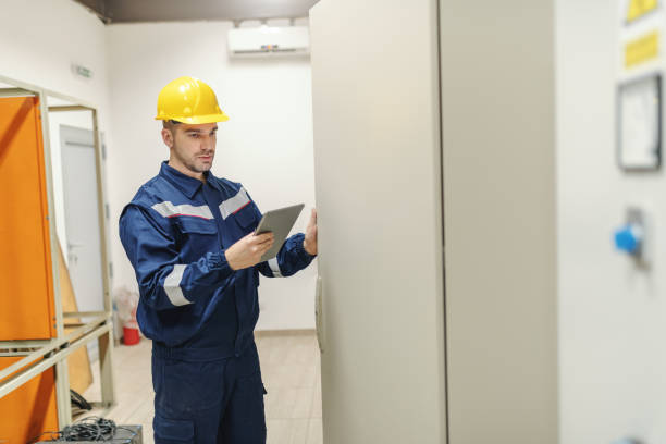 Caucasian dedicated electrician in in uniform and helmet on head using tablet and opening door with dashboard while standing in control room in heavy industry plant.