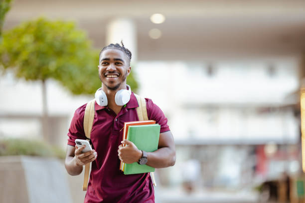 Smiling male student holding books and using smart phone. He is walking and using smart phone