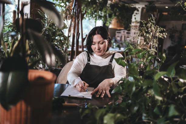 Creative short-haired brunette wearing dark apron sitting at a table in a plant-filled space, concentrating as she writes in her notebook.