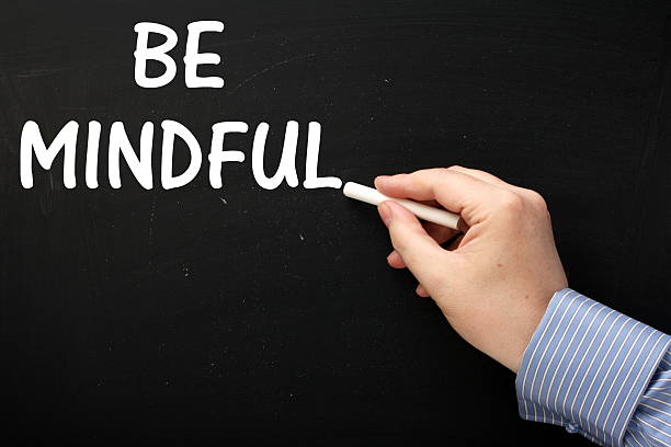 Male hand wearing a business shirt writing the phrase Be Mindful on a blackboard. Being Mindful is a mind and body approach to achieving well-being and reducing stress levels
